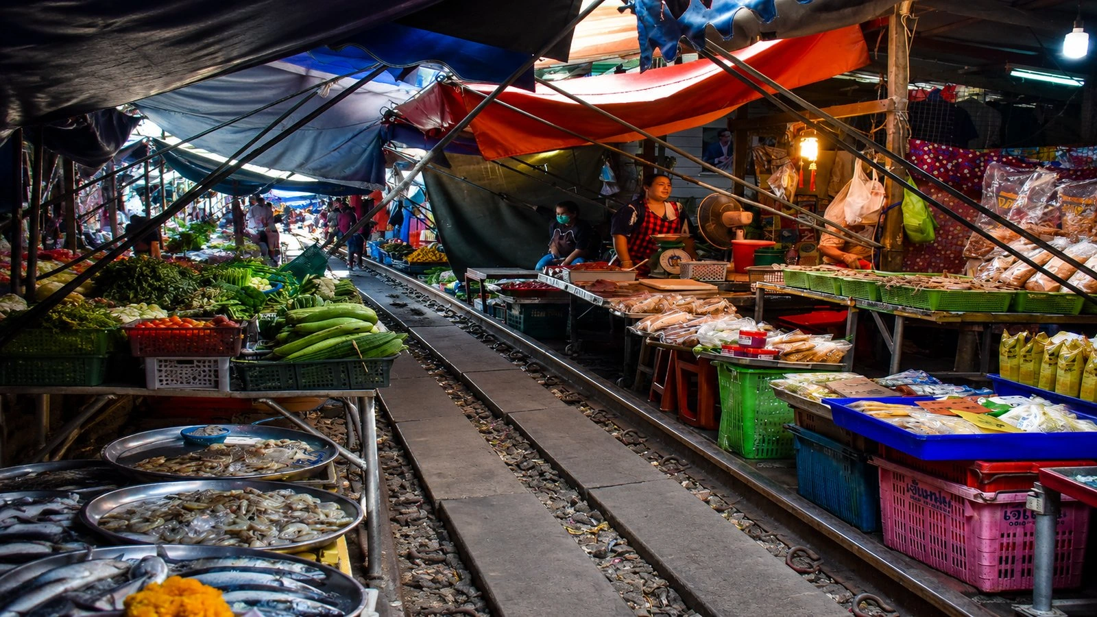 Vendor folding back an awning, fresh seafood and fruit on the rails at Maeklong railway market
